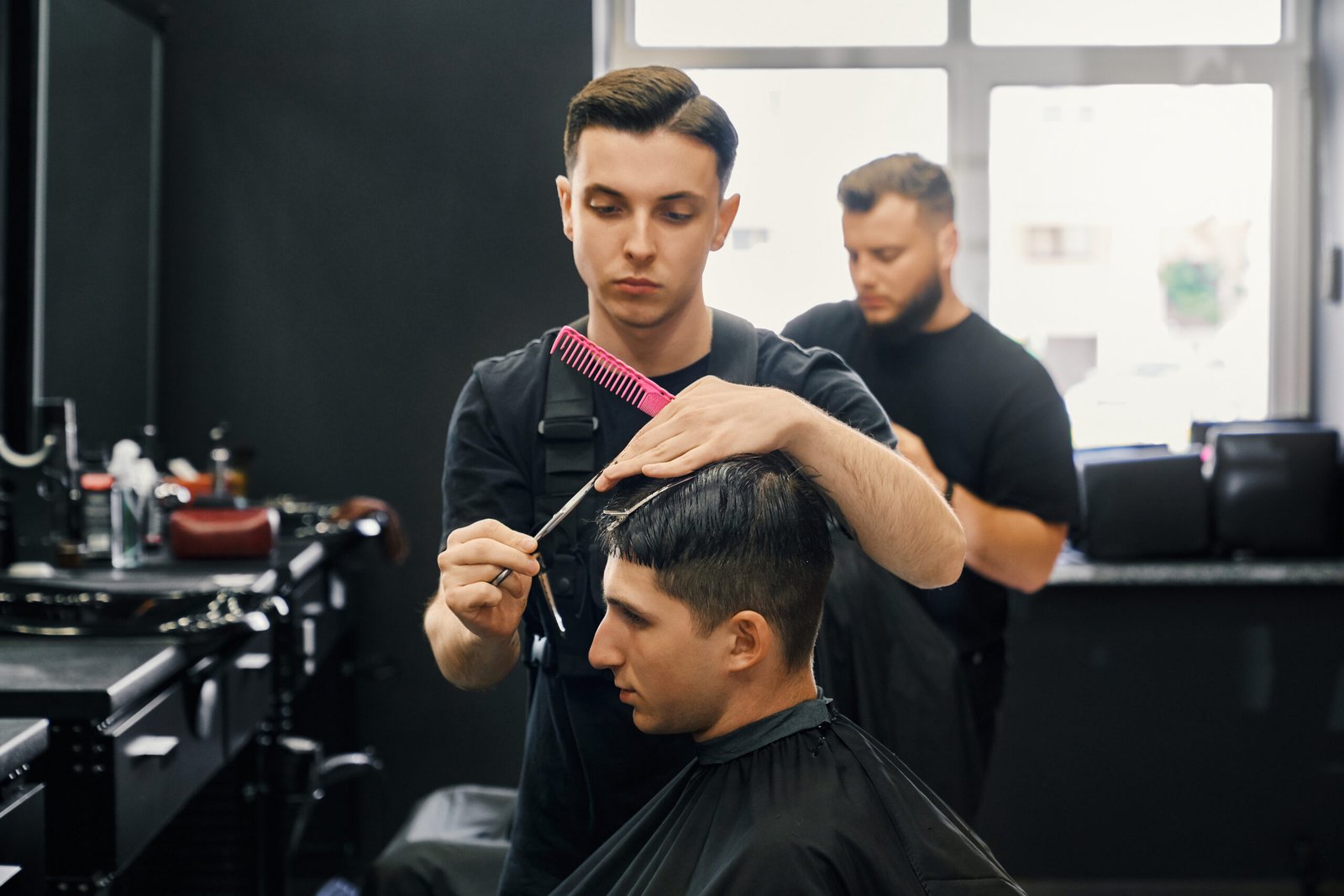 Side view of caucasian male client getting trendy haircut at modern barbershop. Professional barber using scissors and comb while styling hair of young man.